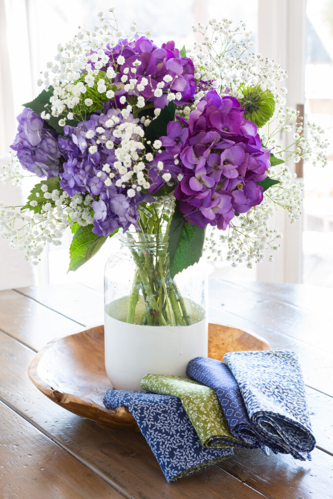 Bottom painted glass vase filled with purple, hydrangeas, white and green flowers. Placed on a wood bowl with handmade cloth napkins folded along the edge.