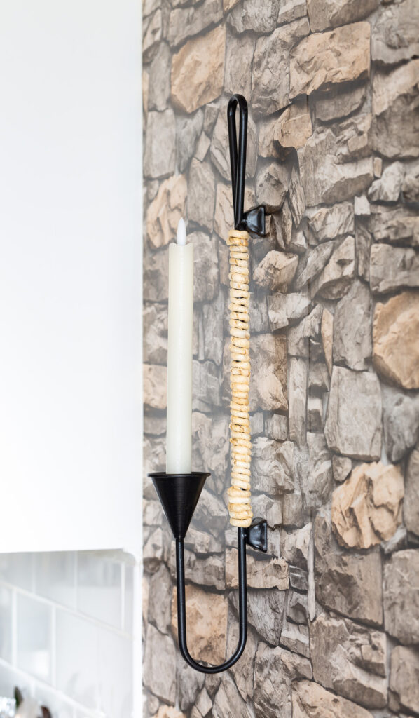 Black metal and natural cord wrapped candle sconce hanging on a wall with stacked stone wallpaper.