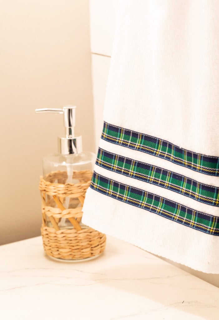 White hand towel with 3 rows of blue and yellow plaid ribbon added to make it more decorative. It is hanging on a chrome towel ring in a bathroom.