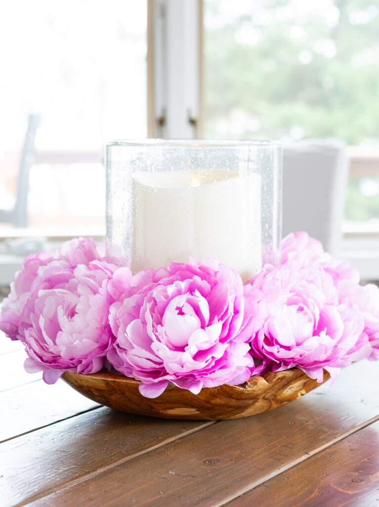 Pink flowers surrounding a large candle that is placed in a large bowl on a wood table.