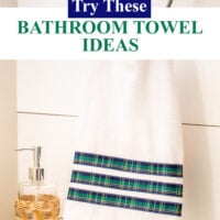 White hand towel with 3 rows of blue and yellow plaid ribbon added to make it more decorative. It is hanging on a chrome towel ring in a bathroom.