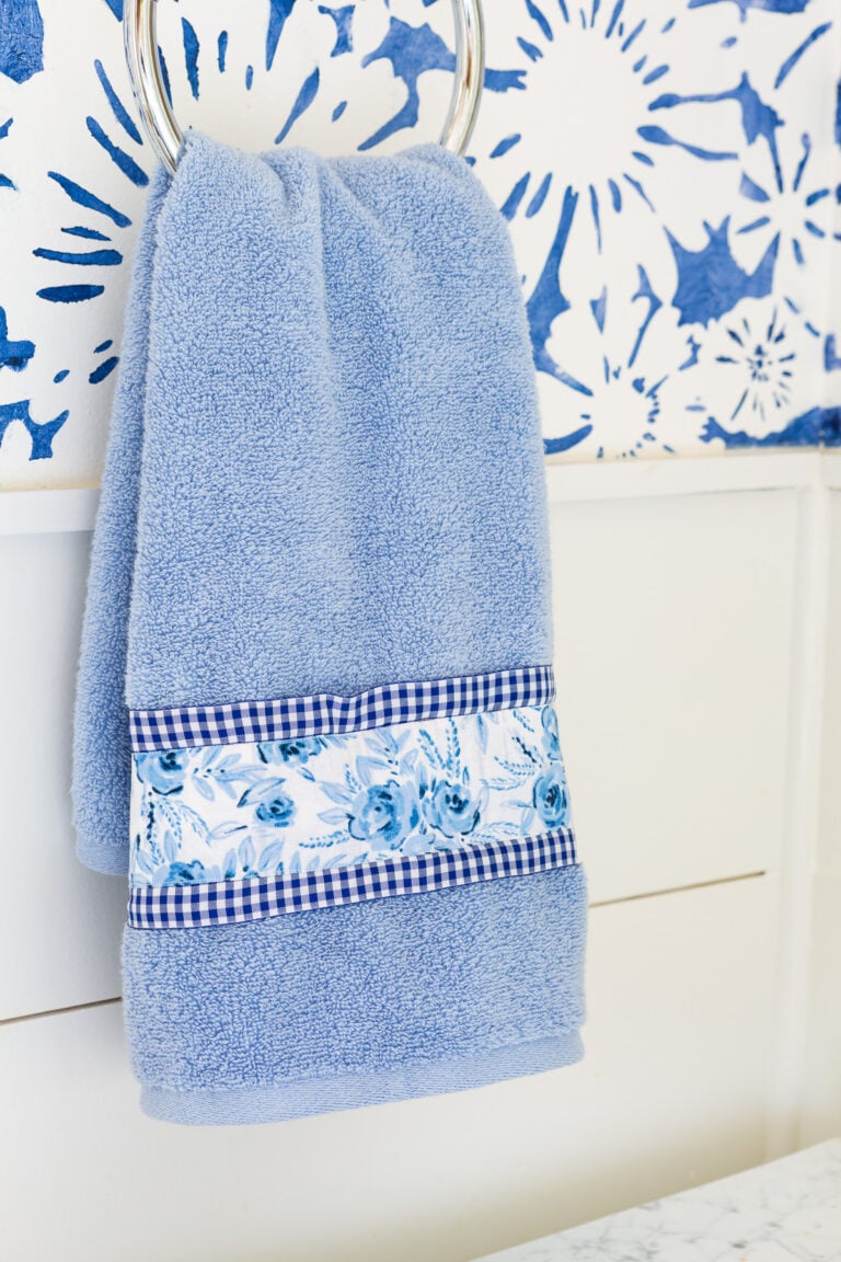 blue hand towel hanging on towel hook in bathroom. Towel is embellished with blue and white fabric and ribbon.