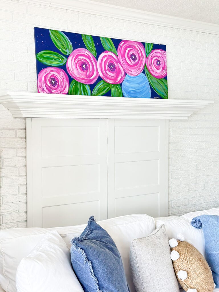 DIY wall art painting of big pink flowers on a dark blue background placed on a living room mantel.