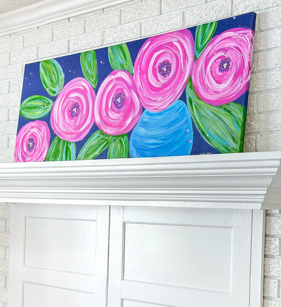 DIY wall art painting of big pink flowers on a dark blue background placed on a living room mantel.