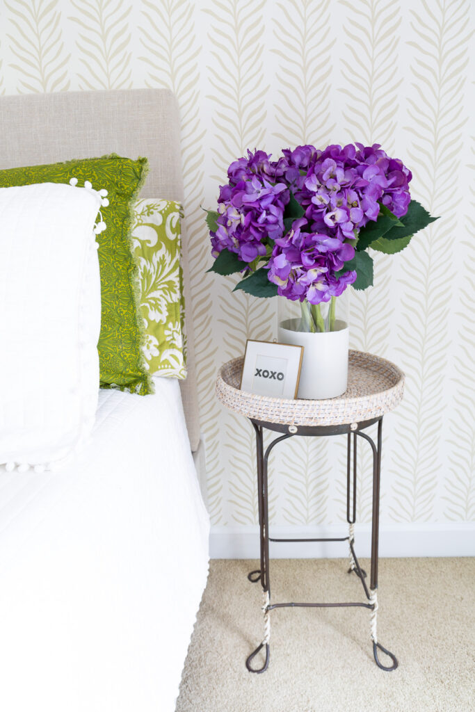 A vase a purple hydrangeas in a glass vase on a table. Neutral beige and white wallpaper on wall behind it from Love vs. Design