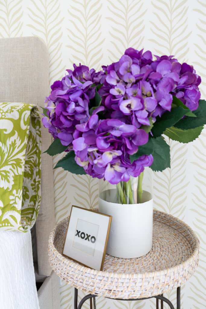 A vase a purple hydrangeas in a glass vase on a table. Neutral beige and white wallpaper on wall behind it from Love vs. Design