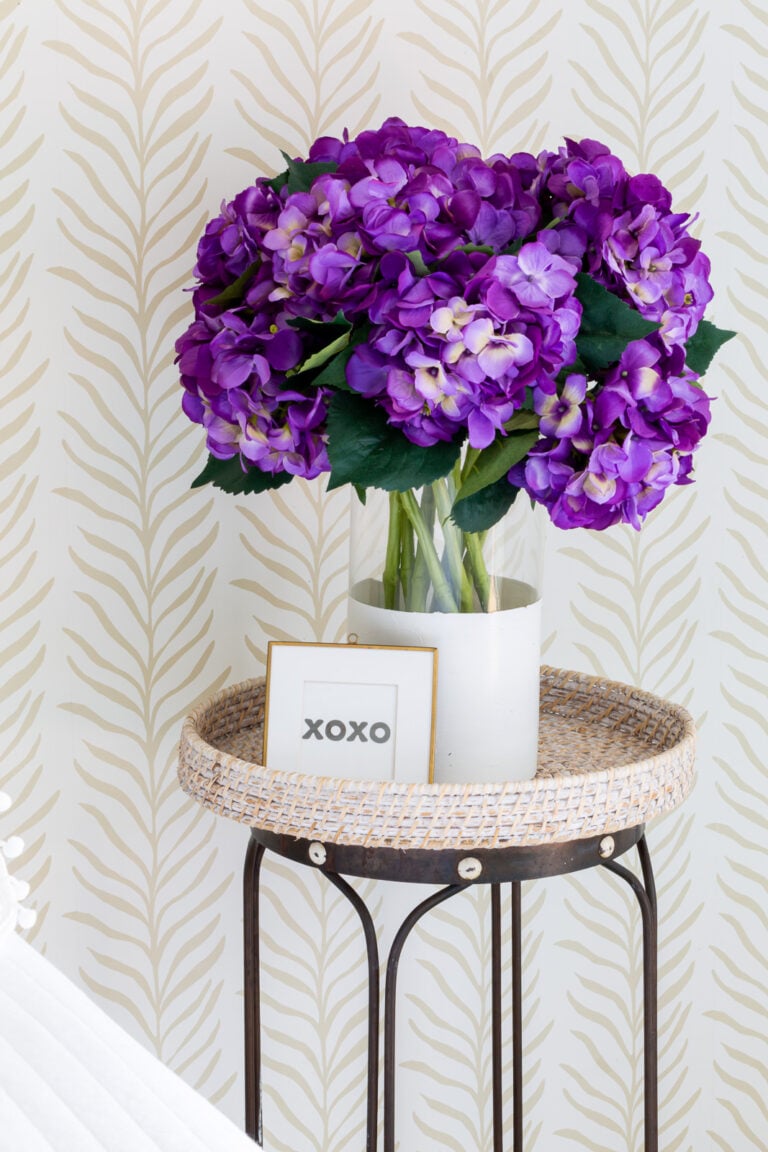 A vase of purple hydrangeas on a side table along a bedroom wall covered with neutral color wallpaper.