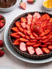 A side shot of vegan chocolate strawberry tart on a white plate with bowls of pastry cream, strawberries and apricot jam.