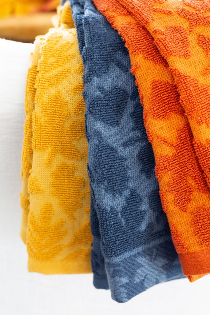 3 fall hued dishtowels with a textured leaves draped over a chair.