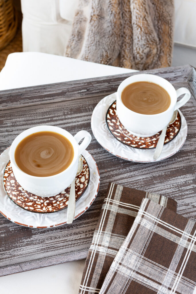two white mugs on a brown wood tray on an ottoman. Mugs are on brown splatterware plates with coffee in them.
