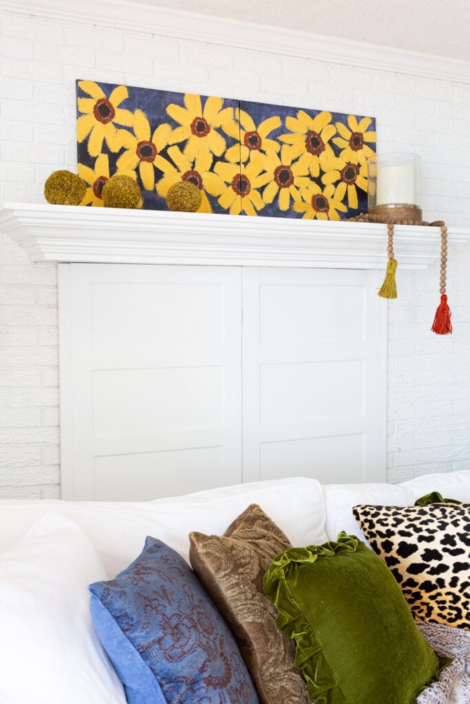 Side view of painted fall flower canvas on a mantel above a white sofa with fall colored and textured throw pillows.