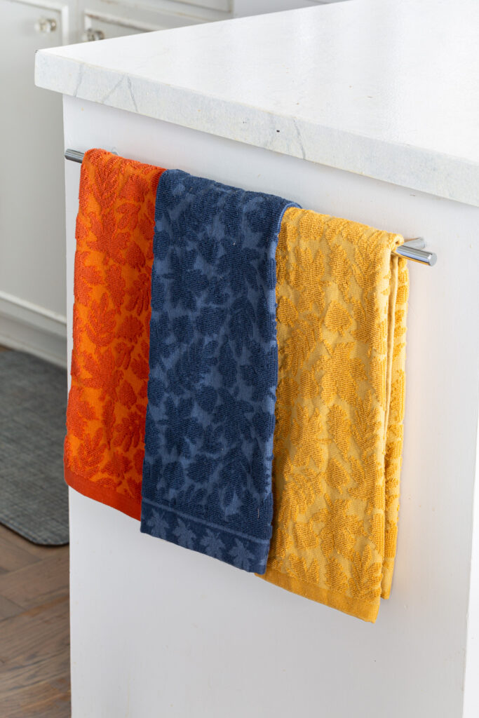 3 fall colored dish towels hanging on a towel bar in a white kitchen.
