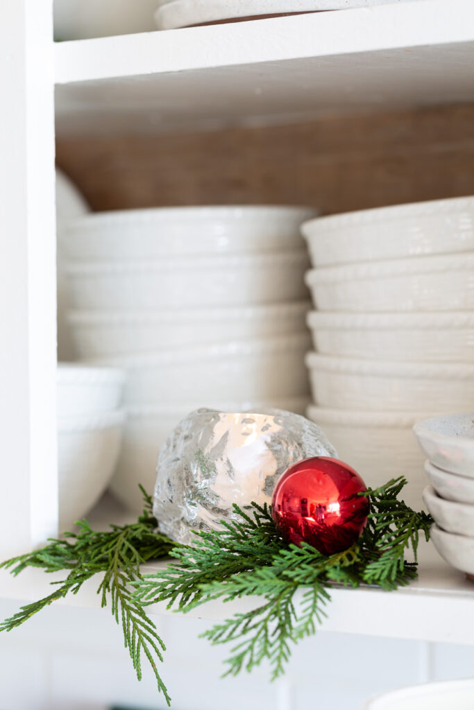 Kosta Boda snowball votive holder on an open kitchen shelf with greenery around it and a red ball ornament.