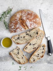 Sliced no-knead olive bread on a grey surface with a bread knife, sprigs of rosemary and a bowl of olive oil.