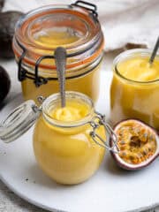 Three jars of vegan passion fruit curd on a plate with a halved passion fruit.