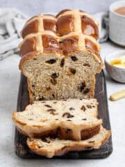 Sliced vegan hot cross bun loaf on a wooden board with a cup of tea and a bowl of butter.