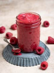 Vegan raspberry curd in a jar on top of a metal dish with a spoon and some fresh raspberries.
