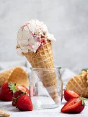 A cone of vegan strawberry cheesecake ice cream in a glass jar surrounded by fresh strawberries and ice cream cones.