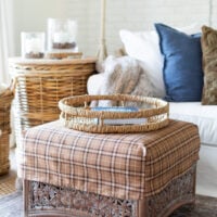 a square rattan ottoman with a brown plaid slipcover on it in from of a white sofa with layers of fall decor on it.