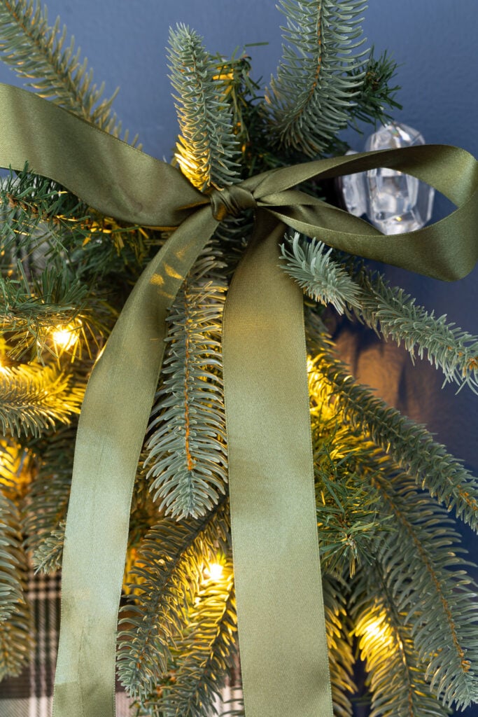 Green satin bow tied onto a King of Christmas Noble Fir garland that is draped on the top of a bed headboard to decorate a bedroom for Christmas.