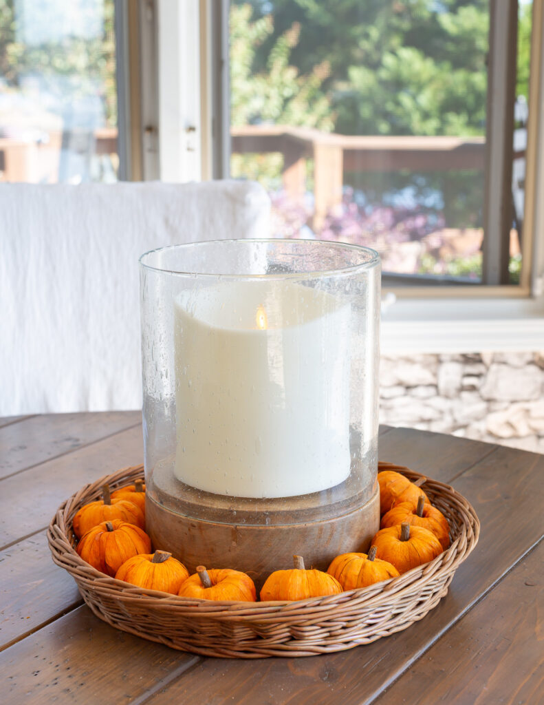 Large white candle in round tray basket with mini-pumpkins around it.