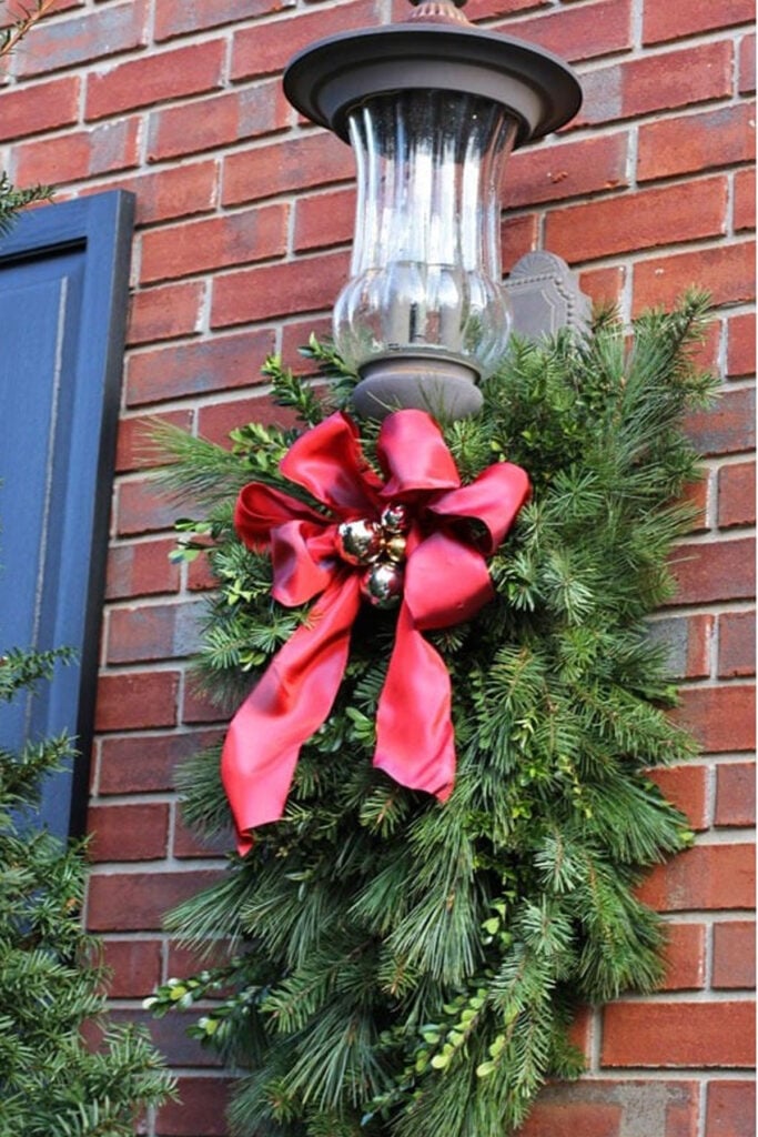 Large front porch light decorated for Christmas using a clever DIY trick to hang greenery and attach ribbon.