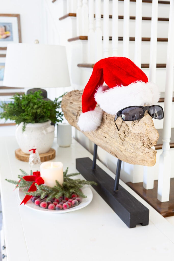 A large decorative piece driftwood modern art topped with a Santa hat and sunglasses on a foyer sideboard as Christmas decor.