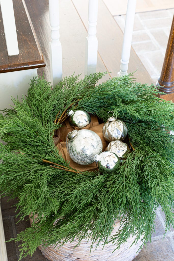 Adding silver Christmas ornaments placed in the center of a greenery wreath on top of a floor basket.